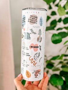 a person holding up a white tumbler with stickers on it and green plants in the background