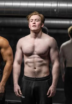 two men standing next to each other in front of a metal wall with no shirt on