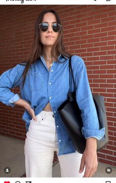 a woman in white pants and blue shirt holding a black handbag while standing next to a brick wall