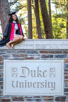 a woman sitting on top of a brick sign in front of trees and wearing a graduation gown