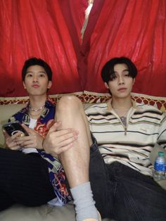 two young men sitting on a couch with their legs crossed and one holding a cell phone