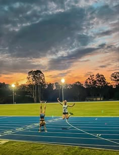 two people jumping up in the air on a blue and green running track at sunset