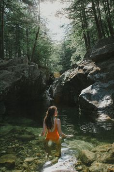 a woman in an orange bathing suit wading through a river surrounded by rocks and trees