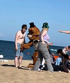 three people standing on the beach next to a giant teddy bear wearing a top hat