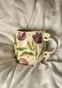 a coffee cup sitting on top of a bed covered in white sheets with purple flowers