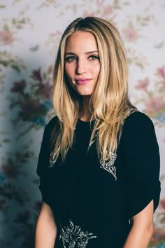 a woman in a black dress standing next to a floral wallpapered wall and smiling at the camera