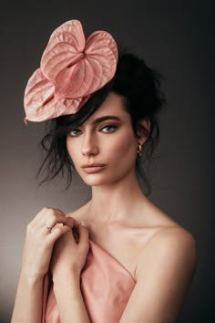 a woman wearing a pink dress with a large flower on top of her head and hands near her chest