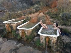 a woman laying in an outdoor bathtub surrounded by rocks and grass, with two large tubs on each side