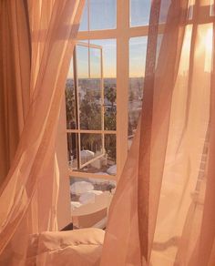 an open window with sheer curtains in front of a cityscape and the sun shining through