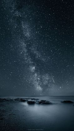 the night sky is filled with stars and some rocks in the water, as well as an airplane flying overhead