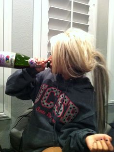 a blonde woman sitting in front of a mirror holding a wine bottle to her ear