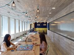 two women sitting at a long table playing board games in a large room with windows