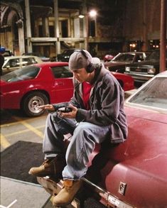 a man sitting on the hood of a red car looking at his cell phone while listening to headphones