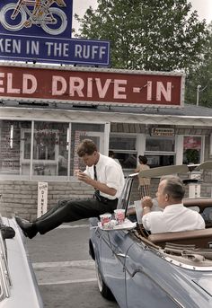 two men sitting in an old car at the drive - in, talking on their cell phones