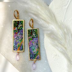 Add a touch of artistic elegance to your look with these stunning handmade earrings inspired by Claude Monet's artist's garden in Giverny painting. These earrings are made from high-quality polymer clay and feature an impressionist style that captures the essence of the original artwork.The earrings are designed with a golden metal rectangular frame that beautifully frames the intricate design. They are also accented with a delicate lavender glass bead that dangles from the bottom, adding a pop Artist Garden, Earrings Handmade Clay, Artsy Earrings, Precious Metal Clay Jewelry, Glasses Inspiration, Jewelry Making Classes, Metal Clay Jewelry, Dope Jewelry, Funky Jewelry