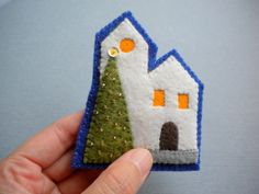 a hand holding a small felt house ornament with a tree in front of it