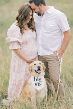 a pregnant couple and their dog pose for a photo in the tall grass at sunset