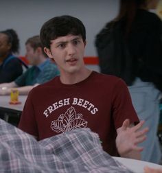 a young man in a red shirt is talking to someone at a table with other people