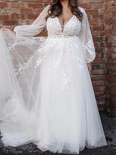 a woman standing in front of a brick wall wearing a white dress with sheer sleeves