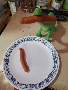 a plate with a hot dog on it and a toy frog holding a carrot in its mouth