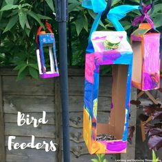 two colorful bird feeders hanging from a pole in front of some trees and bushes