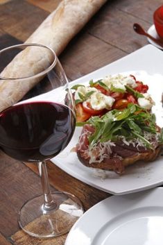 a glass of red wine and some food on a white plate next to bread with tomatoes, cheese and lettuce