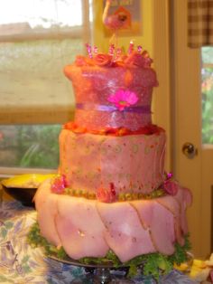 a three tiered cake with pink frosting and flowers on the top, sitting in front of a window