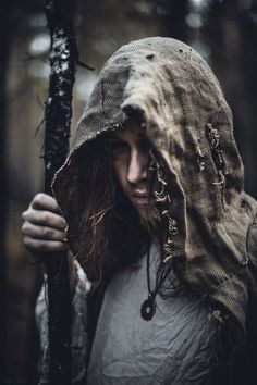 a man with long hair wearing a hooded jacket and holding a stick in the woods