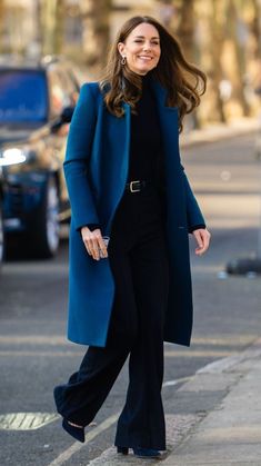 a woman walking down the street wearing a blue coat and black pants with her hands in her pockets