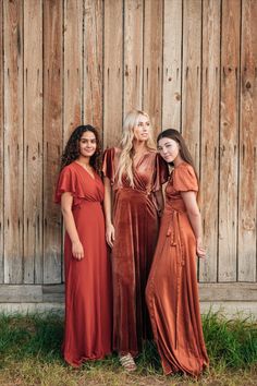 three women standing next to each other in front of a wooden wall wearing long dresses