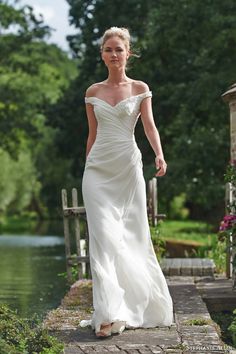 a woman in a white dress is walking on a path by the water and trees