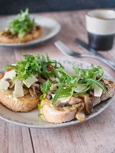 an open face sandwich with mushrooms and greens on a plate next to a cup of coffee