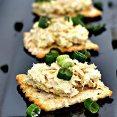 three crackers with various toppings on them sitting on a black surface, one has green onions and the other is topped with cheese