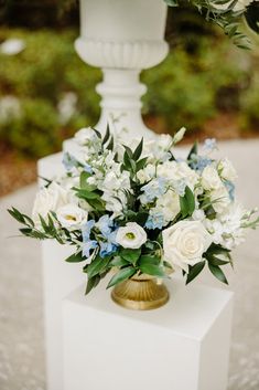 white and blue flowers are in a gold vase on a pedestal at an outdoor ceremony