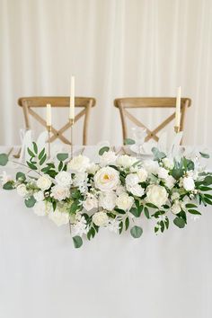 white flowers and greenery are arranged on a table with two chairs in the background