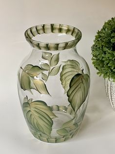 a glass vase with leaves painted on it next to a potted plant in the background