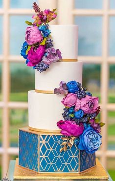 a three tiered wedding cake with flowers on the top and bottom, sitting on a table in front of a window