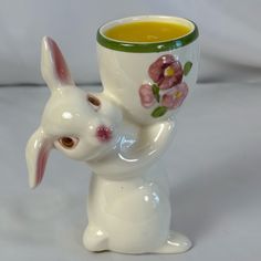 a white ceramic bunny holding a cup with yellow liquid in it's mouth and flowers painted on the side