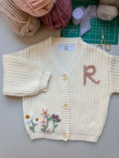 a knitted cardigan with flowers on it next to yarn and crochet hooks