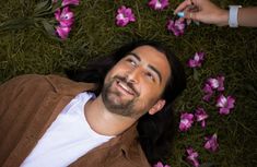 a man laying on the ground surrounded by pink flowers with one hand reaching for something