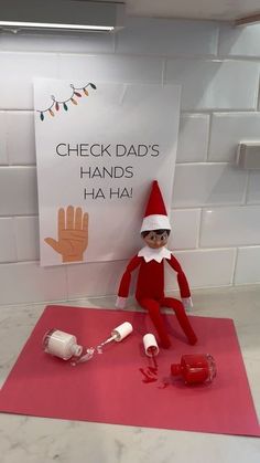 an elf is sitting on top of a pink mat next to a sign that says check dad's hands ha ha ha