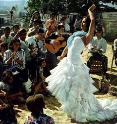 a woman in a white dress is dancing on the beach while people are sitting and standing around her