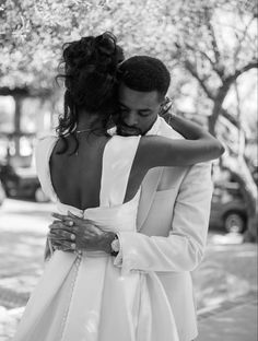 a man and woman embracing each other in front of a tree on the sidewalk with their arms around each other