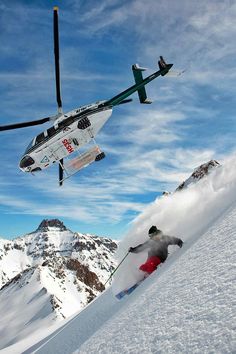 a helicopter flying over a man skiing down a snow covered slope with mountains in the background