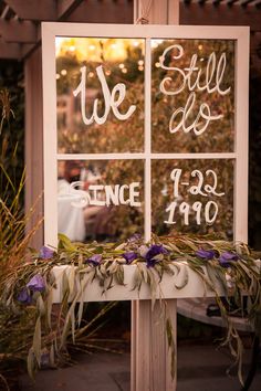 a sign that says we still do and since in front of a window with purple flowers