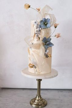 a three tiered white cake with blue flowers on top and gold stand against the wall