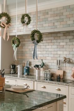 Four small wreaths with ribbons hang above a modern kitchen island with a brick backsplash. Kitchen Cabinet Display, Festive Kitchen, Above Kitchen Cabinets, Countertop Display