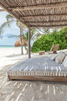 a bed sitting under a thatched roof on top of a sandy beach next to the ocean