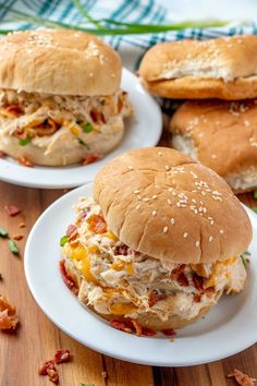 two chicken sandwiches with cheese and bacon on buns sitting on plates next to each other