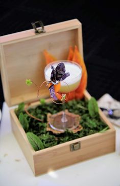 an empty glass in a wooden box filled with green plants and oranges sitting on top of a table